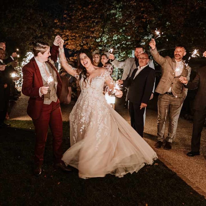 Bride in maroon bridal suit with grey waistcoat, dusky pink tie and autumnal dried flower boutonniere dancing with bride in wearing long sleeve lace illusion sleeve wedding dress with overlay during sparkler exit 
