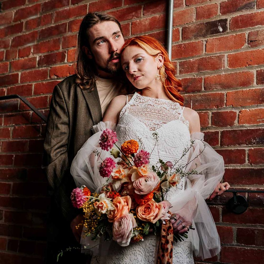 Bride in 70s inspired boatneck short lace wedding dress with detachable tulle puff sleeves holding large colourful mixed flower bridal bouquet with groom in brown blazer, white shirt, grey suit trousers and vans shoes 