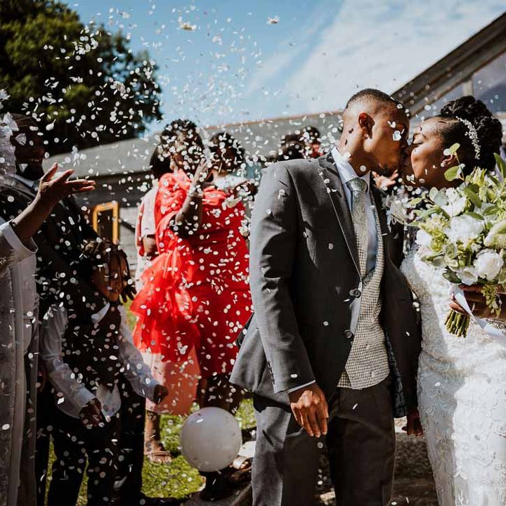 Groom in charcoal grey grooms suit, light grey patterned waistcoat, light grey patterned tie and large white rose and foliage boutonniere kissing bride in lace and mesh boatneck wedding dress with silver bridal hair accessories holding large white rose and foliage bridal bouquet in the midst of biodegradable wedding confetti exit 