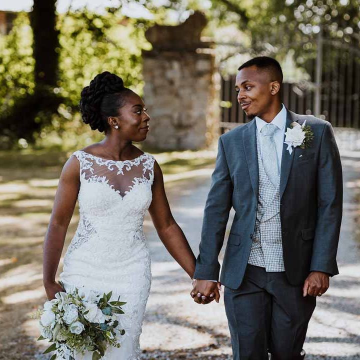Groom in charcoal grey grooms suit, light grey patterned waistcoat, light grey patterned tie and large white rose and foliage boutonniere walking on the grounds of Tredudwell Manor with bride in lace and mesh boatneck wedding dress with silver bridal hair accessories holding large white rose and foliage bridal bouquet 