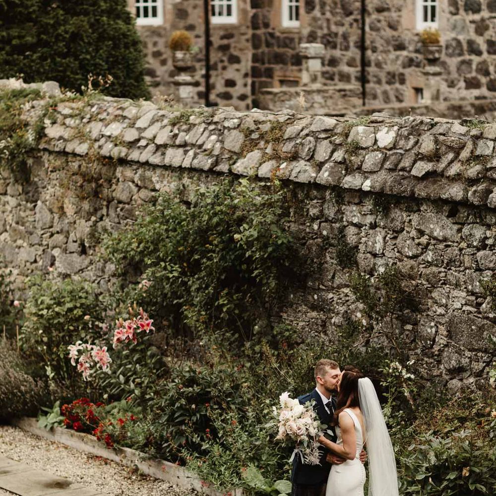 Bride in satin slip wedding dress with church length veil kissing groom in classic black grooms tuxedo at Barra Castle wedding venue Scotland 