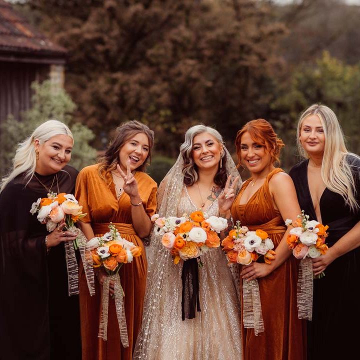 Bride in sleeveless deep v neck sparkly gold wedding dress with sheer and gold polka dot wedding veil standing with bridesmaids in rust and black mismatched bridesmaid dresses