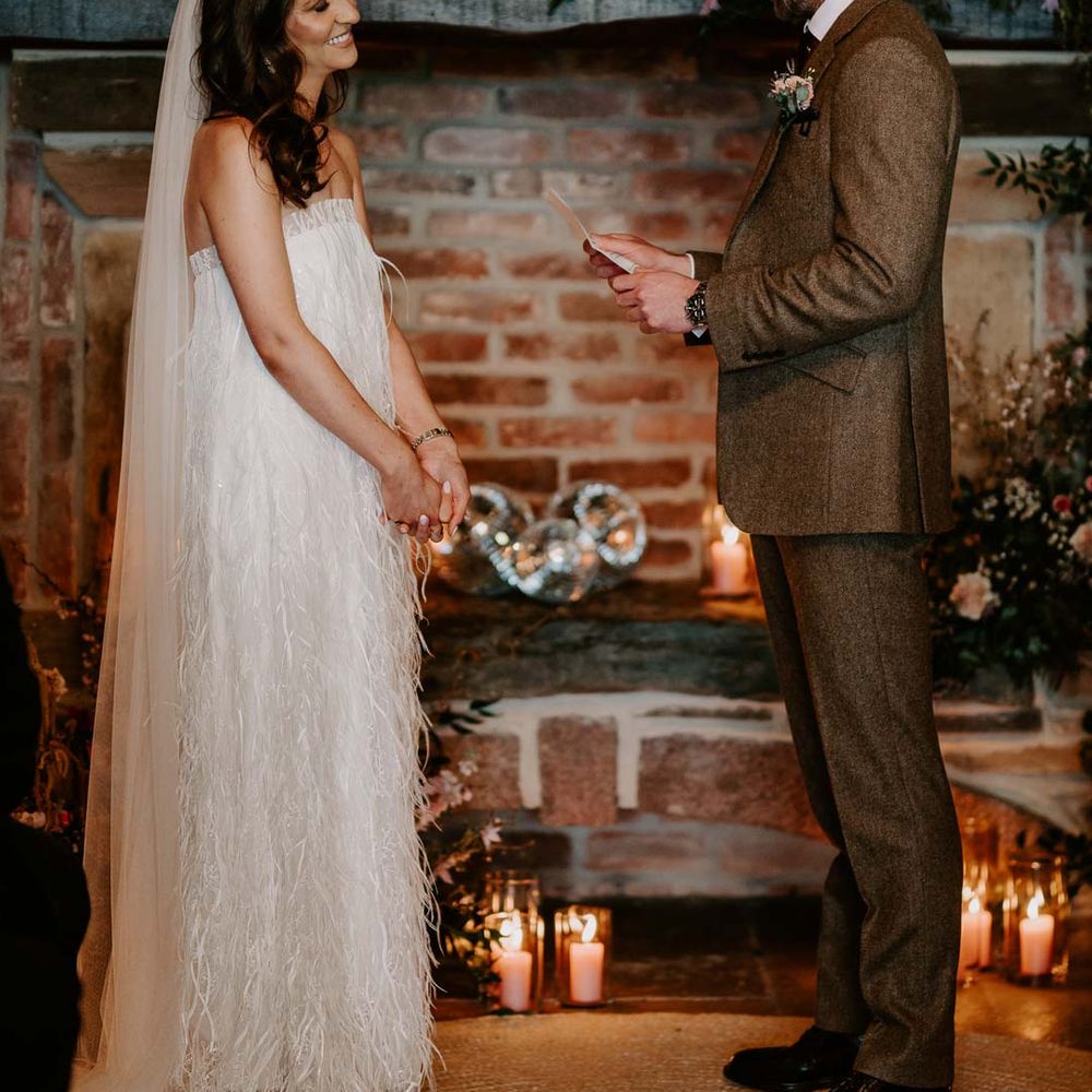 Groom in chocolate brown grooms suit doing wedding reading for bride in strapless ivory fringed crepe gown and chapel length veil standing by large fireplace and disco ball wedding decor