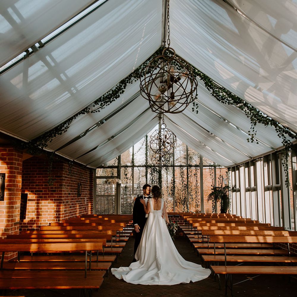 The bride and groom stand in the glasshouse decorated with white drapery and foliage 