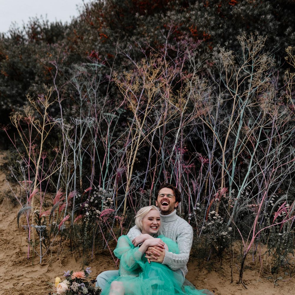 Groom in a grey jumper sitting in the sand embracing his bride in a sheer pastel wedding dress for intimate beach elopement 