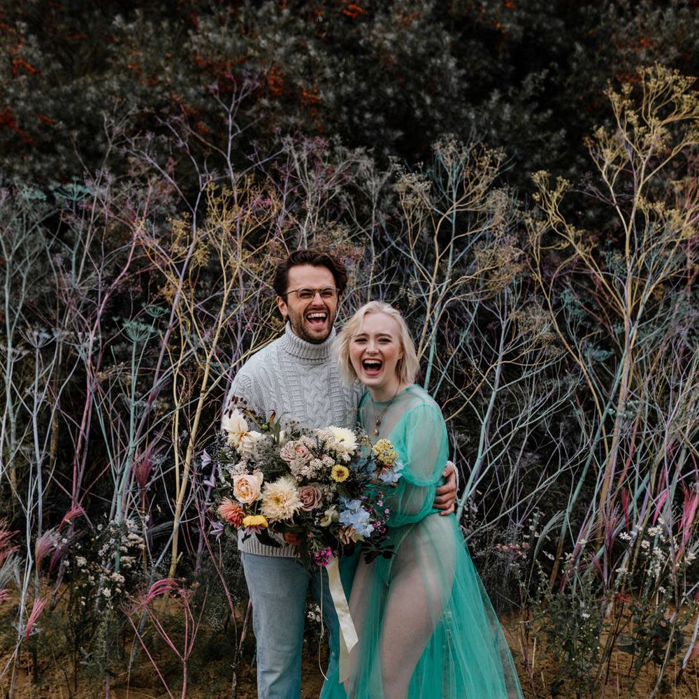 White blonde bride with bob length hair laughing on the beach in a sheer pastel wedding dress with her bearded groom in a grey sweater