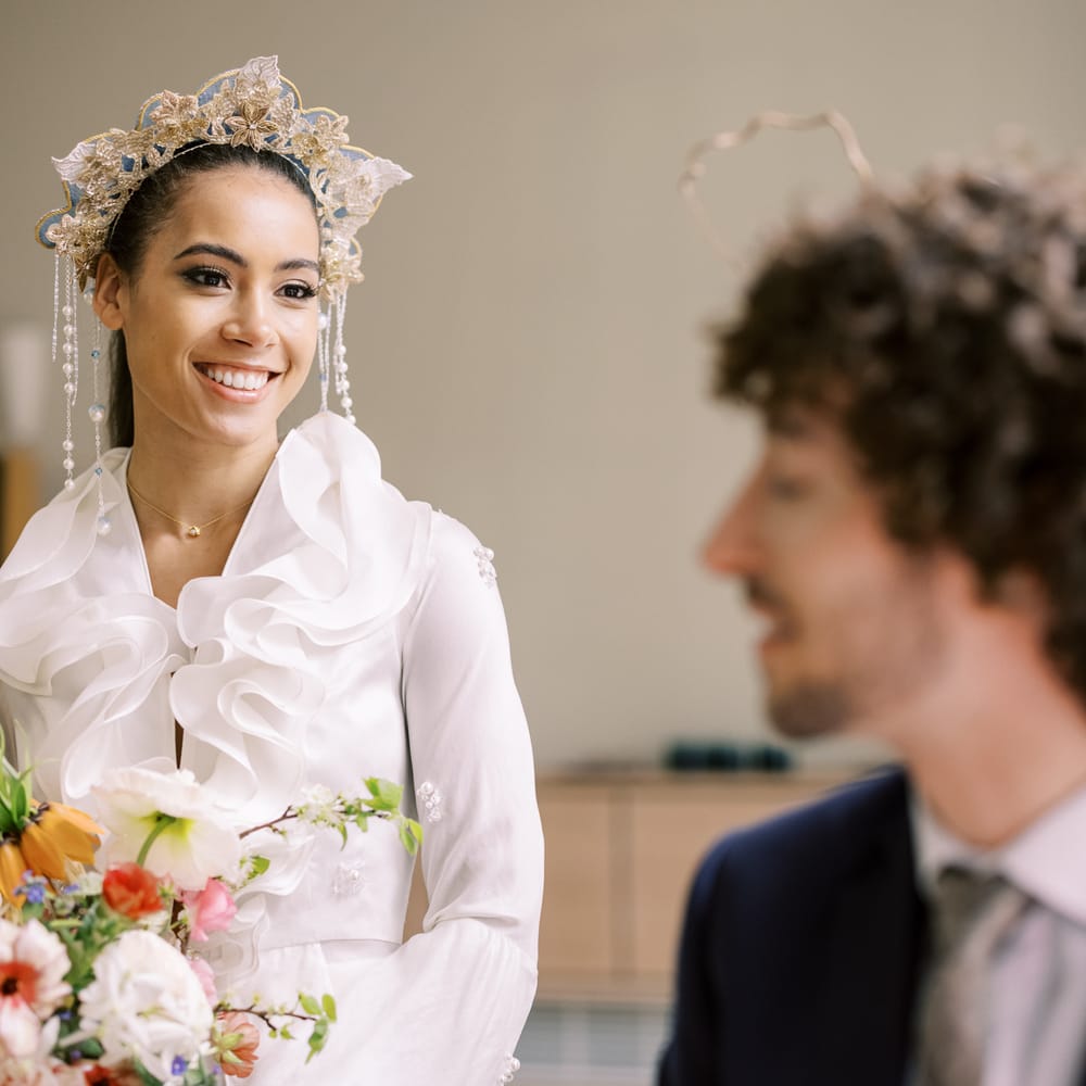 Bride smiles sweetly at groom whilst wearing golden embroidered crown and holding pastel floral bouquet