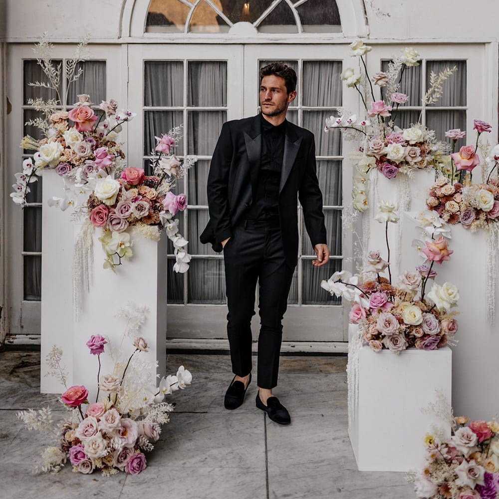 Stylish groom in a black shirt, loafers and tuxedo standing next to a display of romantic pink, lilac white and peach peeled back roses 