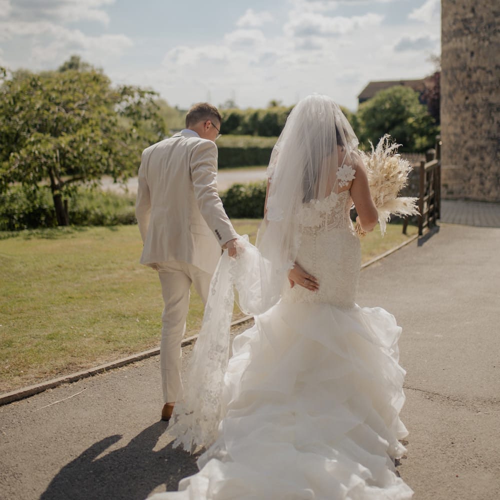 Groom in a beige suit holding his brides appliqué wedding veil as she walks along in her fishtail wedding dress