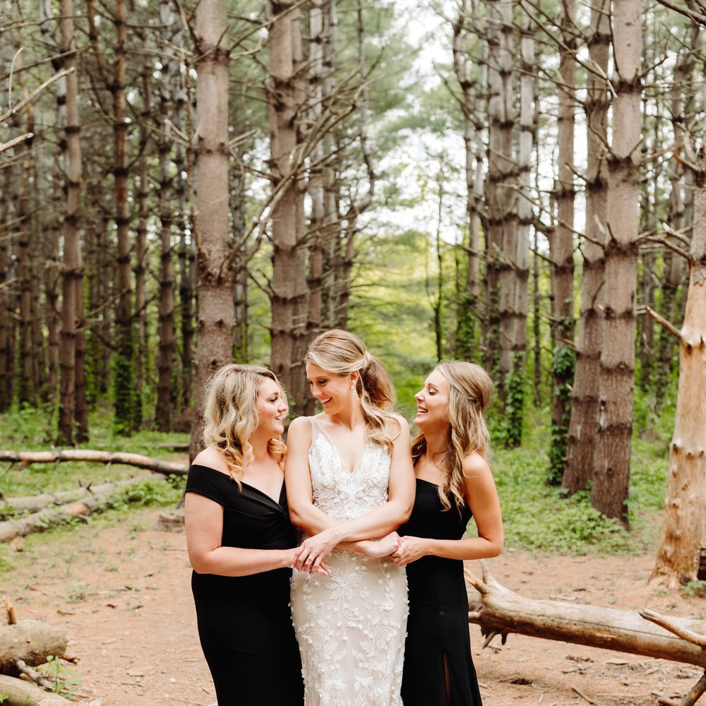 Bride in white embroidered Made With Love wedding dress with train stands in woodland with two bridesmaids in black bridesmaids dresses standing either side of her at backyard wedding