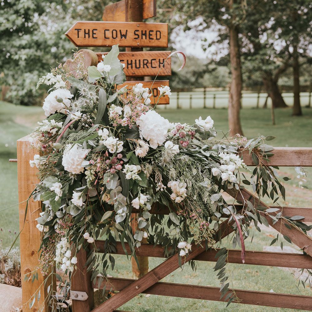 White, pink and green florals and foliage draped over wooden gate next to wooden post sign at Tythe Barn wedding with barn wedding flowers