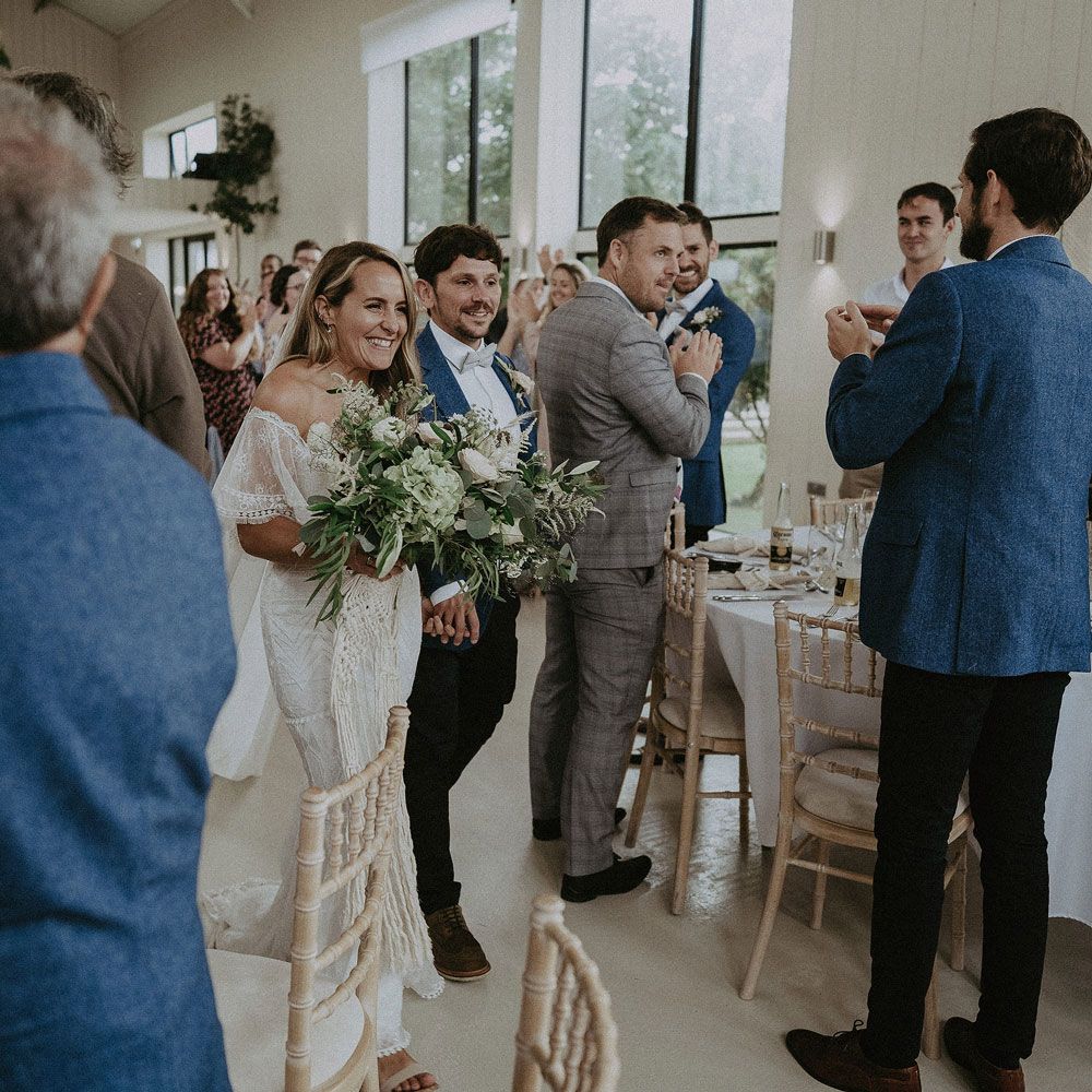 Bride in Grace Loves Lace bardot wedding dress with white and green bridal bouquet walks with groom in blue suit jacket through wedding breakfast at Isle fo Wight wedding with macrame wedding decor