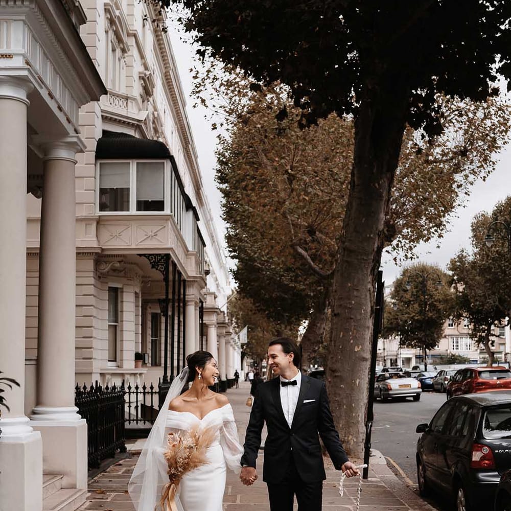 Bride wearing silk fitted wedding dress with cross over detail holding Groom in a black tux with a white shirt walking with dog in a wedding outfit 