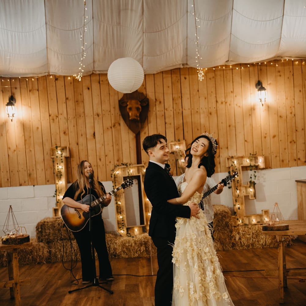 Bride in applique wedding dress and headpiece dances with groom in dark suit whilst guitarists play for rustic wedding reception