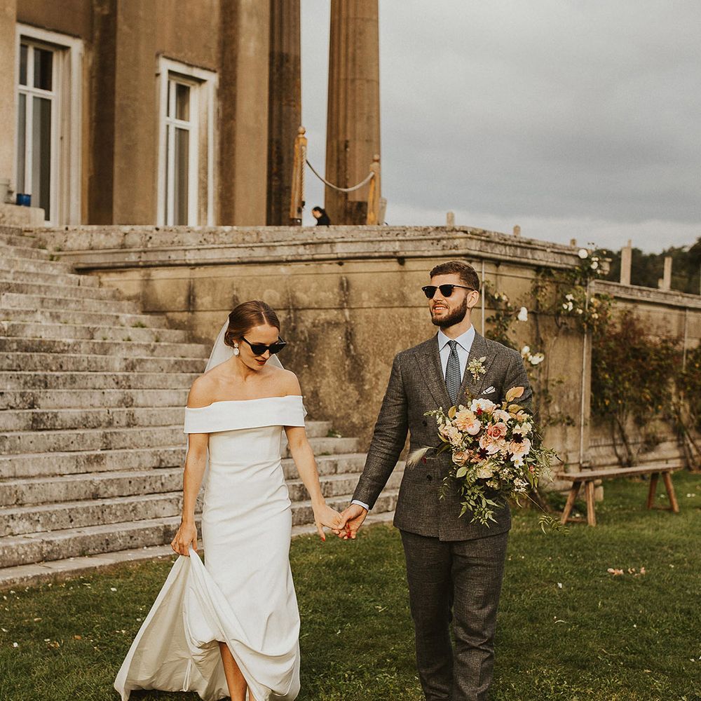 Bride & groom walk hand in hand across lawn on wedding day