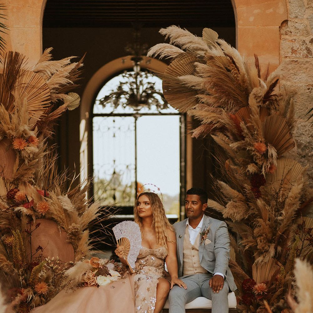 Bride & groom sit together on their wedding day in front of large floral arrangement 