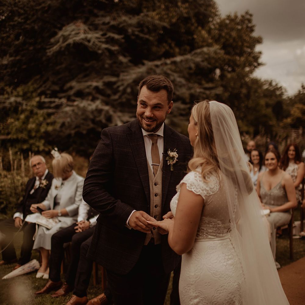 Bride in a lace cap sleeve wedding dress and cathedral length veil exchanging vows with her husband at Dewsall Court