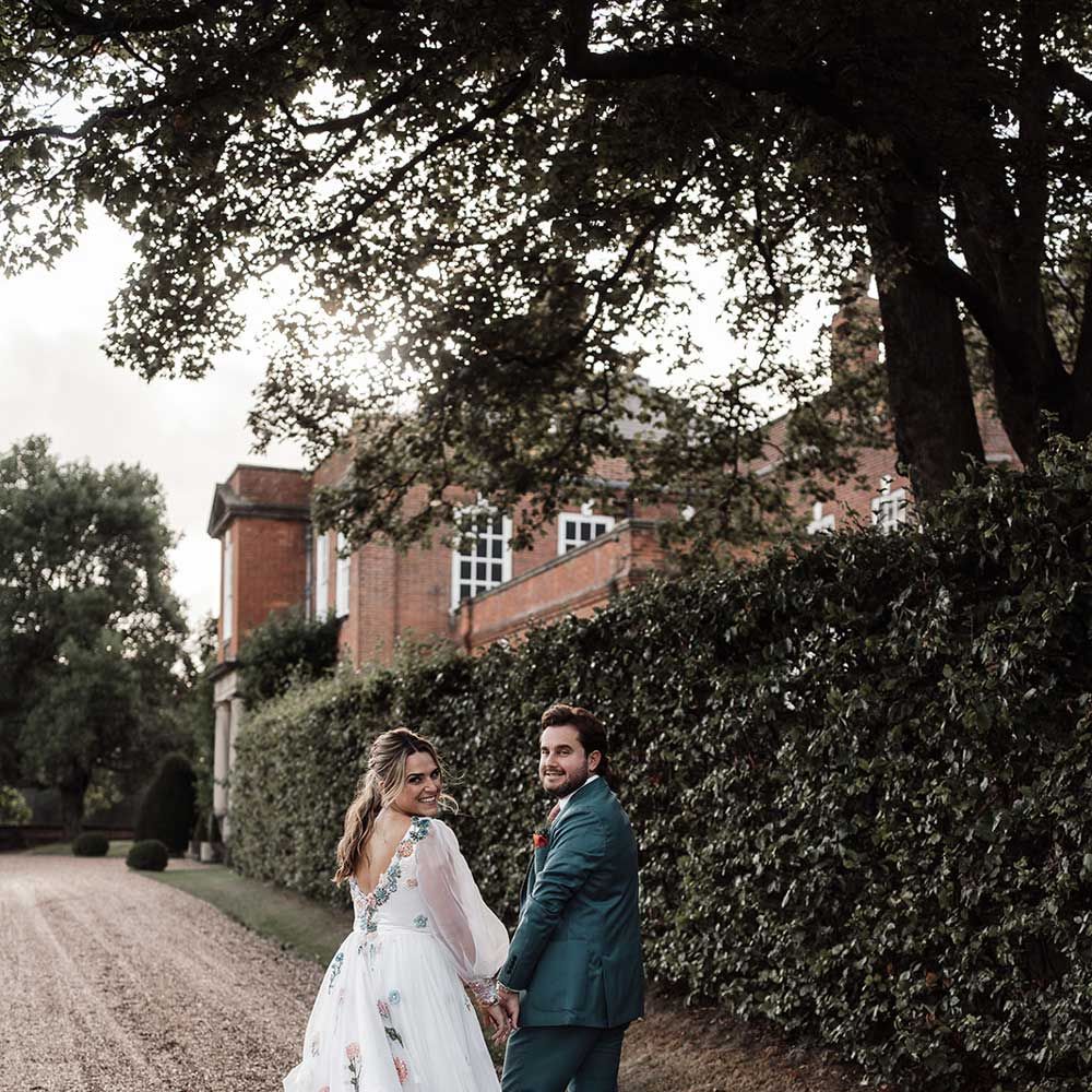 Smiling bride in bespoke Victoria Sanders wedding dress with train and floral embroidery walks down drive holding hands with groom in blue suit