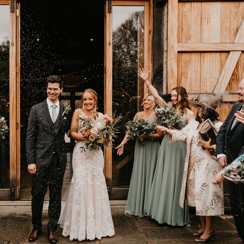 Guests throw confetti over bride in lace and tulle wedding dress holding mixed bridal bouquet and groom in dark suit and blue tie after barn wedding ceremony