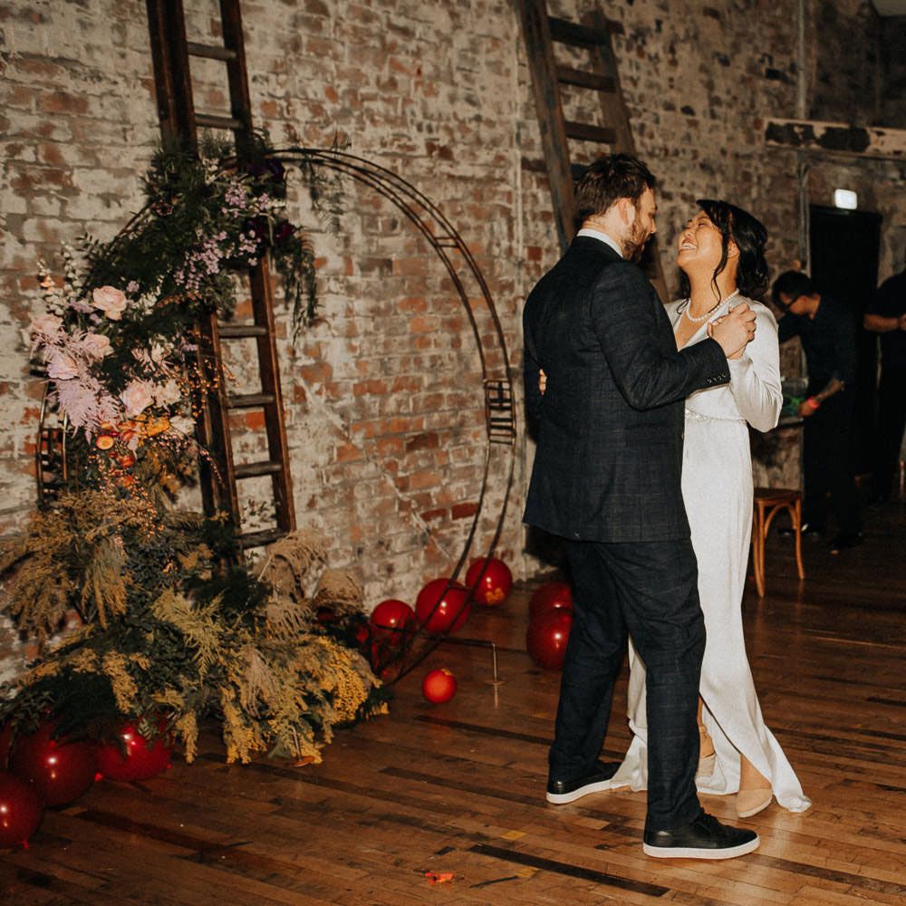 Bride in white long sleeve satin dress and wedges dances with groom in check suit at evening wedding reception