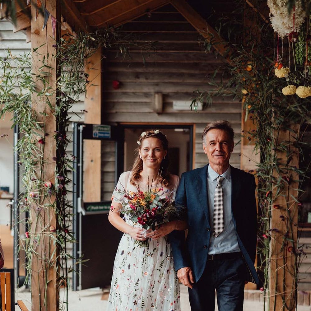 Father of the bride in blue suit and checkered tie walks the bride down the aisle in floral dress
