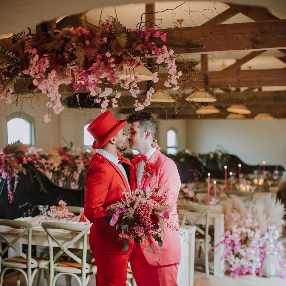 Groom in a red suit and top hat holding a pink flower and dried grass bouquet kissing his partner in a pink suit at their rustic luxe wedding reception at Prestwold Hall Barn 