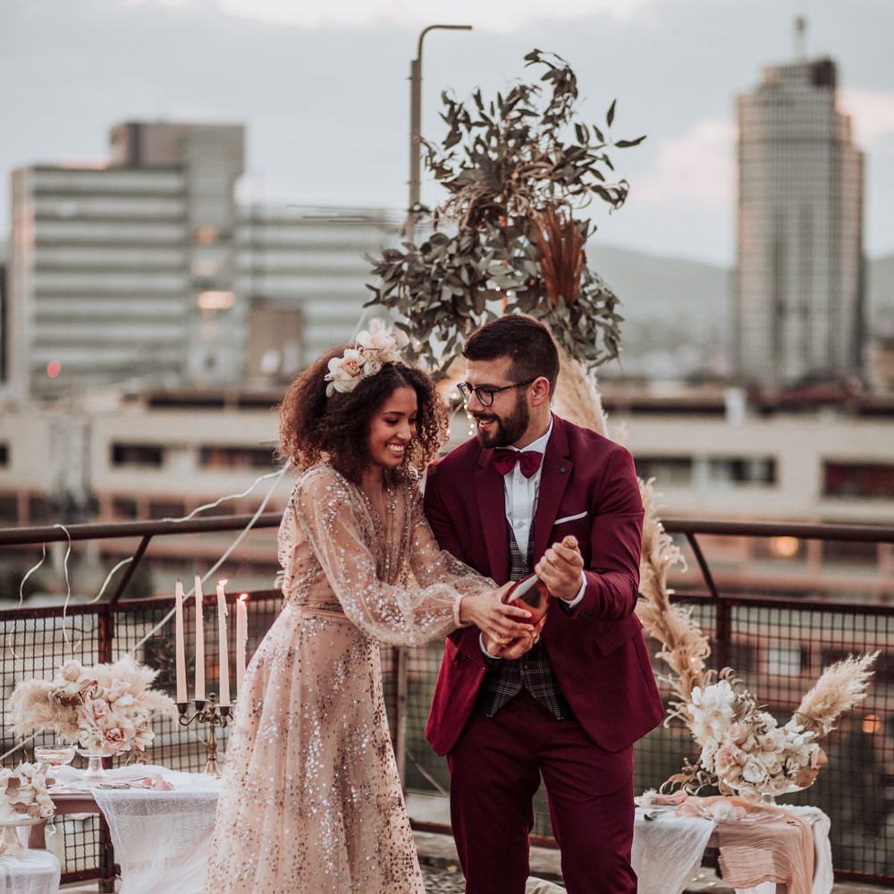 Bride in sparkle wedding dress and groom in maroon suit doing champagne pop