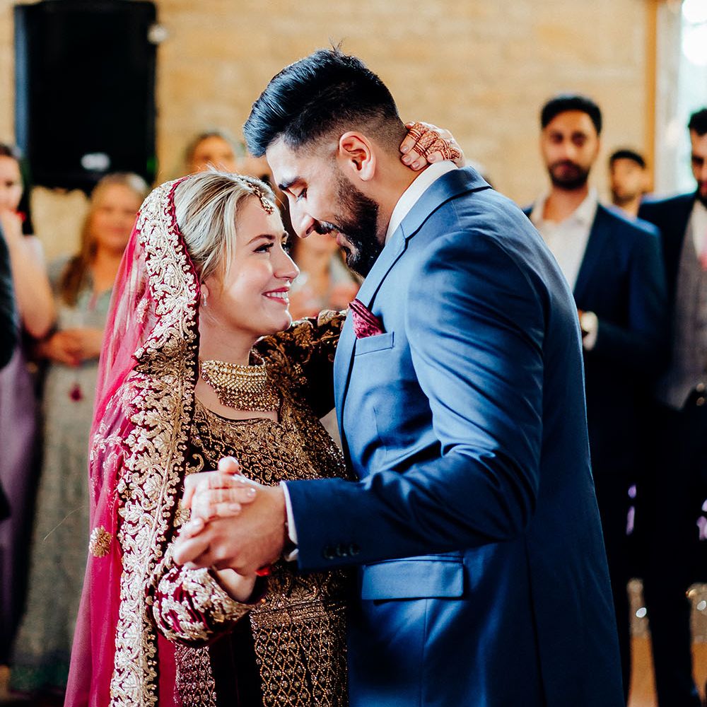 Groom wearing deep blue suit and purple pocket square dances with his bride who wears brightly coloured saree finished with gold embellishment 