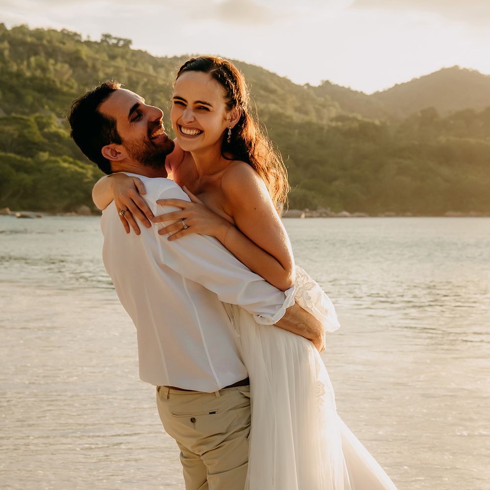 Groom in crisp white shirt, light coloured chinos, tan leather belt and white trainers picking up bride in off the shoulder lace wedding dress with sweetheart neckline and lace floral details on beach in Seychelles