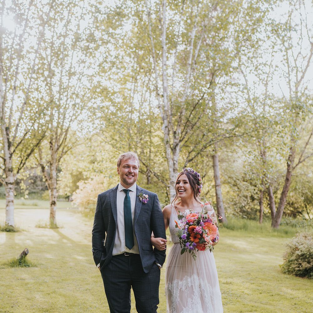 Bride carries colourful floral bouquet as she walks alongside her groom wearing striped suit and colourful floral buttonhole 