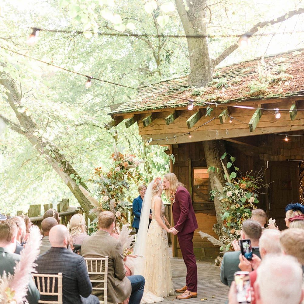 Groom in maroon suit kisses his bride outdoors at the Alnwick Garden Treehouse 
