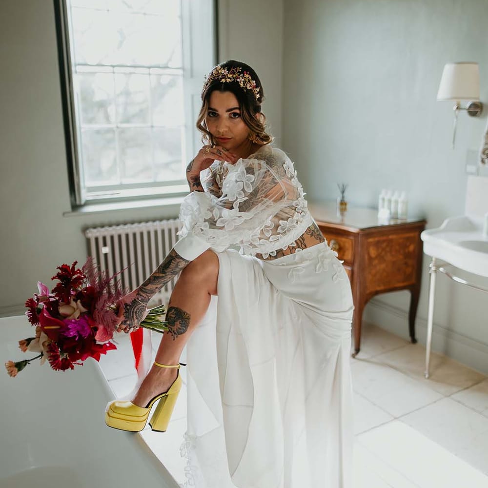 Bride holding hot pink, bright pink and magenta bridal bouquet with dahlias, orchids, roses, anthuriums and dried flowers wearing neon yellow closed toe high heels and two piece bridal separates with puff sleeve sheer crop top with 3d appliqué flowers and full bodied silk skirt and gold bridal flower crown 
