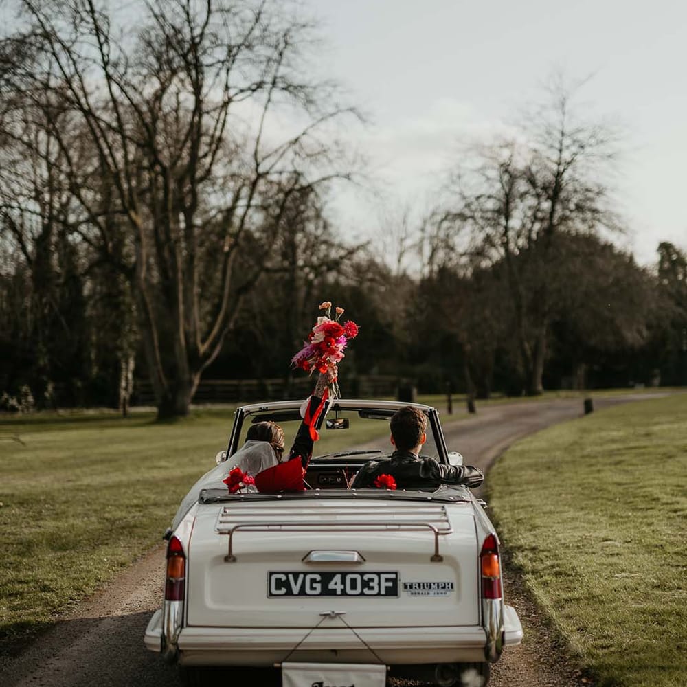Bride holding up hot pink, bright pink and magenta bridal bouquet with dahlias, orchids, roses, amaryllis, anthuriums and dried flowers driving off in off white classic convertible wedding car with white fabric “just married”  banner decorations 