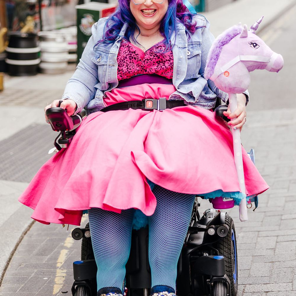 Bride in wheelchair wearing personalised denim jacket, pink wedding dress and blue fishnet tights, gemstone bridal crown, holding fluffy purple unicorn toy