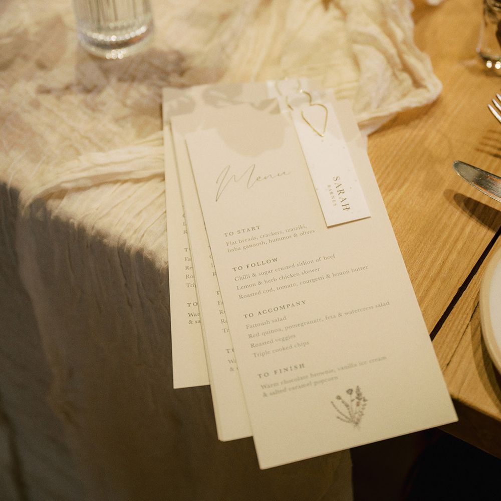 White wedding menu with gold paperclip and white place name on white table runner with small glass vase with white flower arrangements 