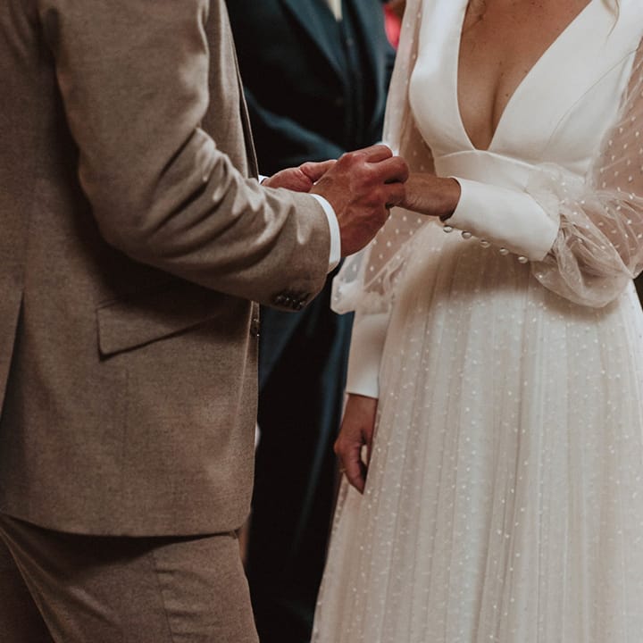 The groom in a neutral coloured suit stands at the altar with the bride and puts on her wedding ring, she wears a Sassi Holford wedding dress with polka dot sleeves