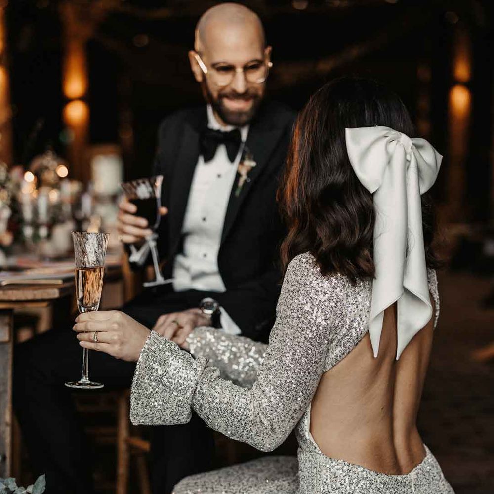 Bride in open back detail long sleeve silver sparkly wedding dress and large white satin bow bridal hair accessory talking to groom in classic sleek tuxedo with black bowtie and boutonniere 