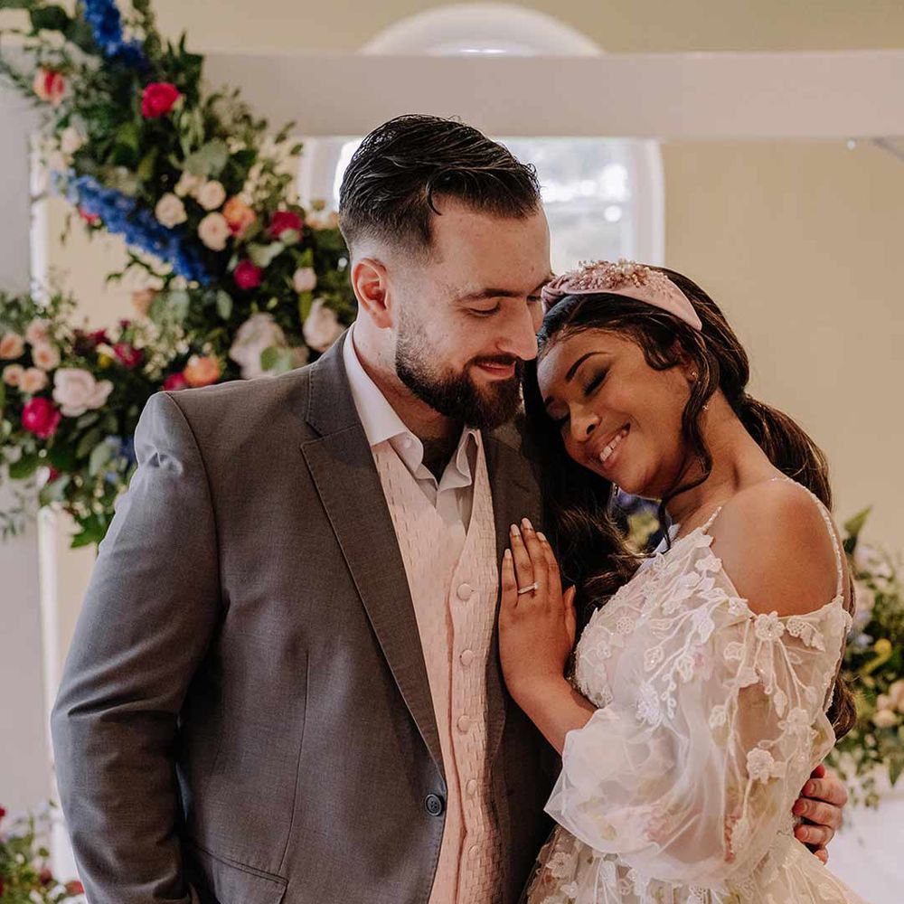 Groom in dark grey suit with cream waistcoat hugging bride in v-neck long sleeve shoulder cut out 3d appliqué wedding dress with flecks of gold wearing satin bridal headband 