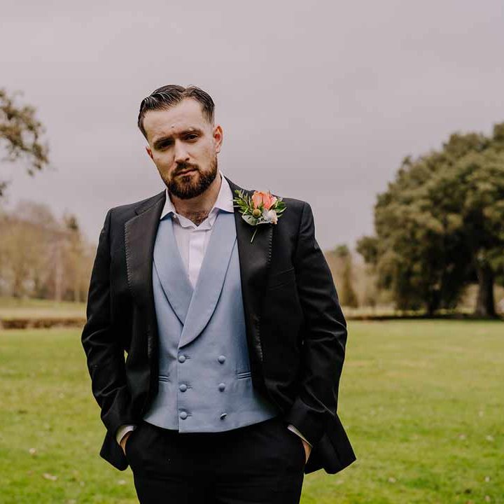 Groom wearing black blazer with pink garden rose and foliage boutonniere, cornflower blue waistcoat and black suit trousers at Braxted Park wedding venue 