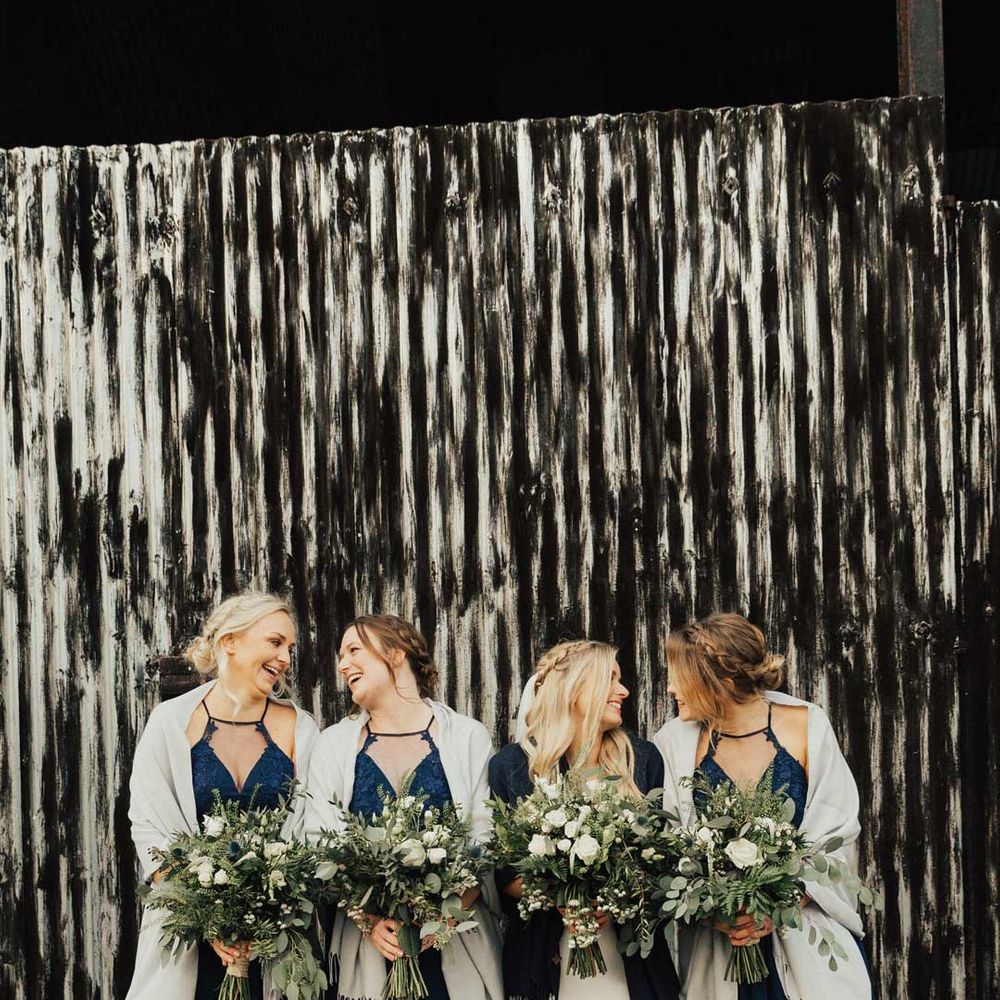 Bride in wedding dress with black shawl standing with bridesmaids in strappy navy bridesmaid dresses and white shawls 