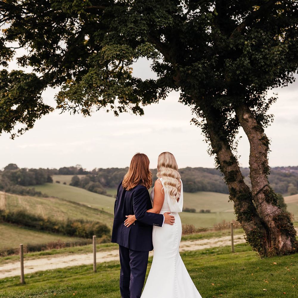 Groom in blue suit looking out at the view of the Surrey Hills with the bride in a draped back Savin London wedding dress with Hollywood curled hair 