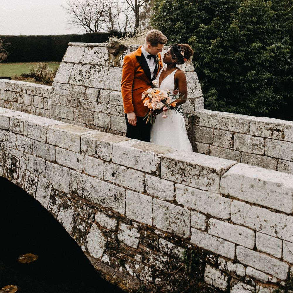 Groom in crushed burnt orange velvet grooms blazer, black bowtie and black suit trousers embracing bride in v neck sleeveless wedding dress with 3D floral applique train holding bridal bouquet on bridge at Brinsop Court wedding venue