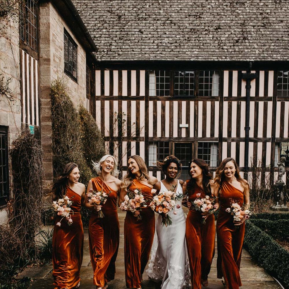 Bridesmaids in one shoulder burnt orange bridesmaid dresses walking with bride wearing v neck sleeveless wedding dress with 3D floral applique train and holding blush and orange garden roses, poppies and carnations, dried flowers and eucalyptus wedding bouquets outside Brinsop Court wedding