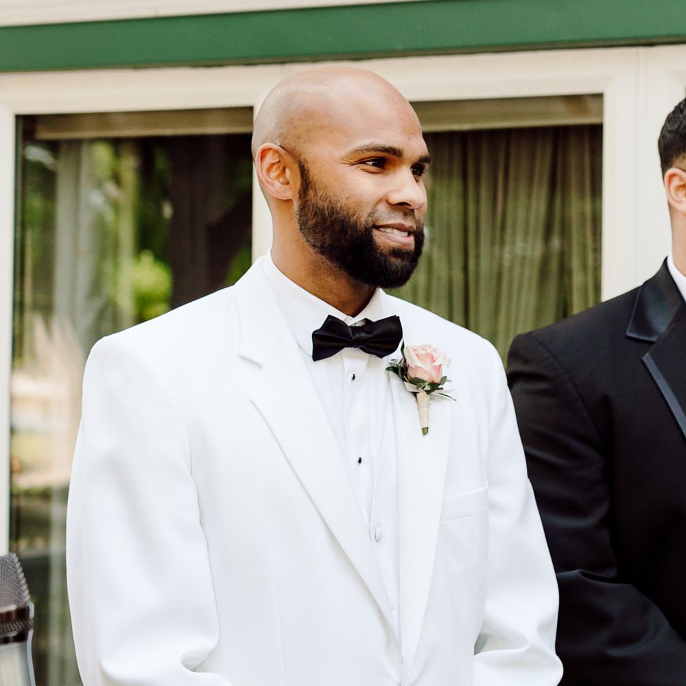 Smiling groom in white shirt, white tuxedo jacket and pink rose buttonhole waits for bride with hands clasped together in front of him at backyard wedding