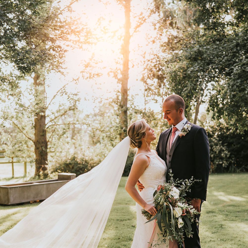 Bride in white lace Justin Alexander wedding dress with train and veil holding white, pink and green bridal bouquet stands with groom in dark three piece suit with pink tie in the gardens at Tythe Barn wedding with barn wedding flowers