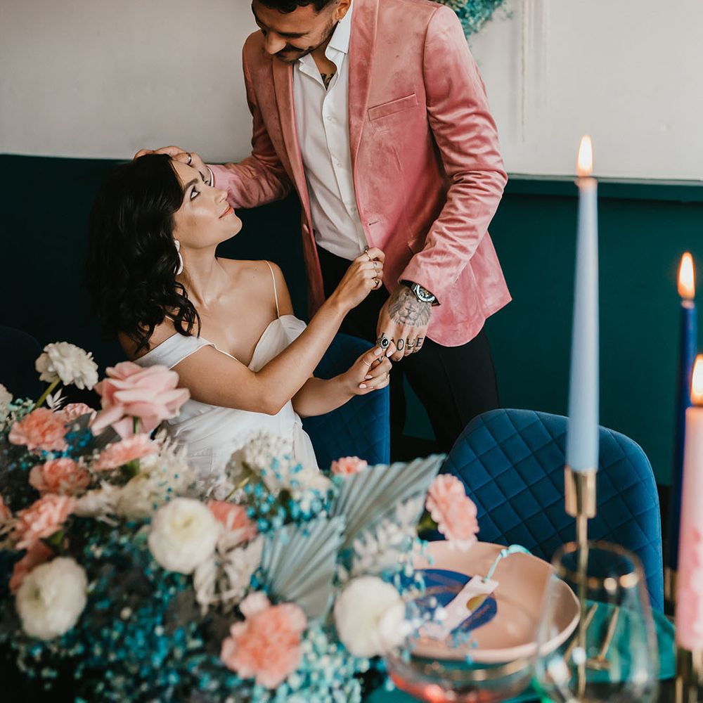 Groom in a pink velvet jacket standing next to his bride in a cold shoulder wedding dress at their stylish pink and teal wedding reception 
