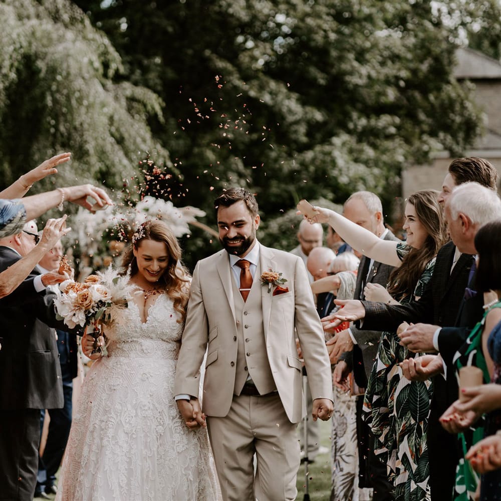Bride in homemade lace wedding dress and veil holding rose bridal bouquet walks holding hands with groom in beige three piece suit whilst guests throw confetti during DIY garden wedding ceremony