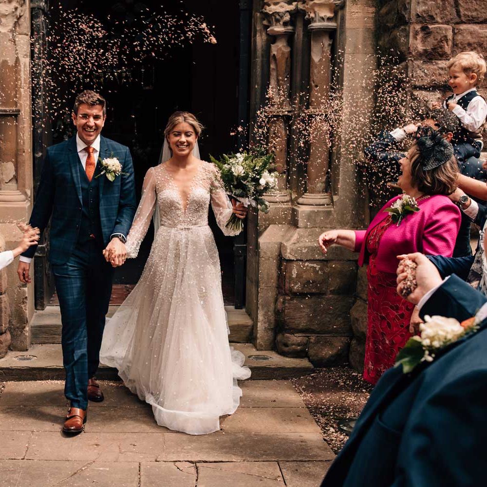 Groom in blue checked three piece suit and tie walks out of church with bride in long sleeve embroidered wedding dress and veil as guests throw confetti