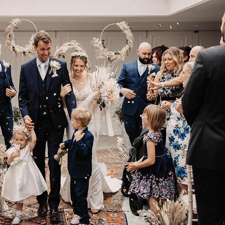 Bride & groom walk down the aisle with their children as wedding guests throw confetti around them