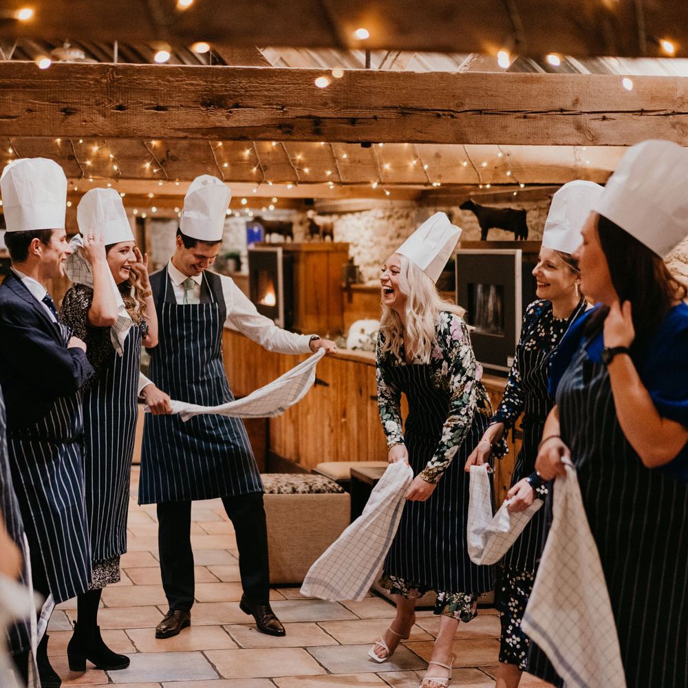 Groom and wedding guests in striped aprons and chefs hats stand in barn for rustic wedding reception